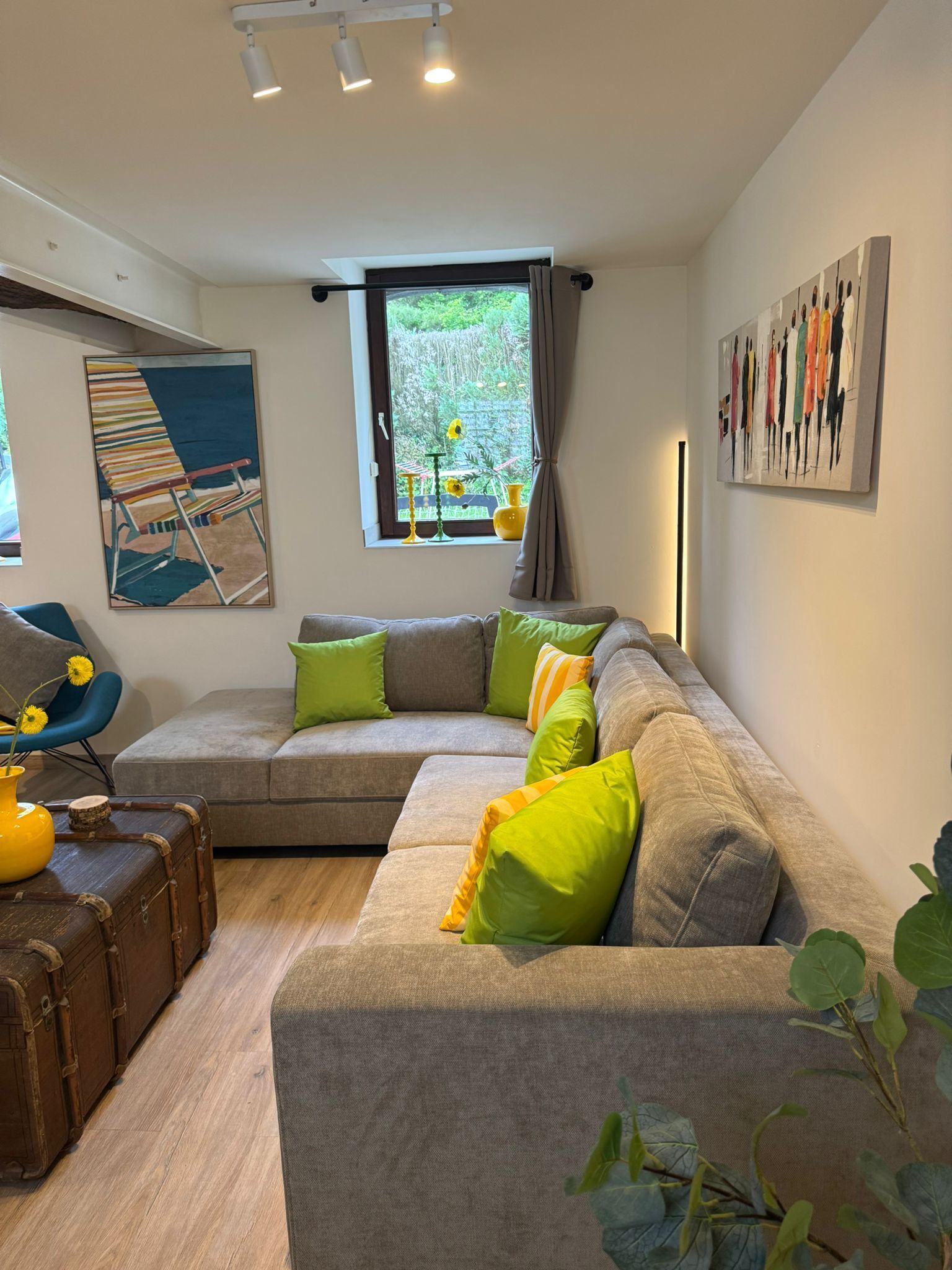 A modern living room with a sectional sofa, a blue chair, a small table with a vase, and a window with curtains.