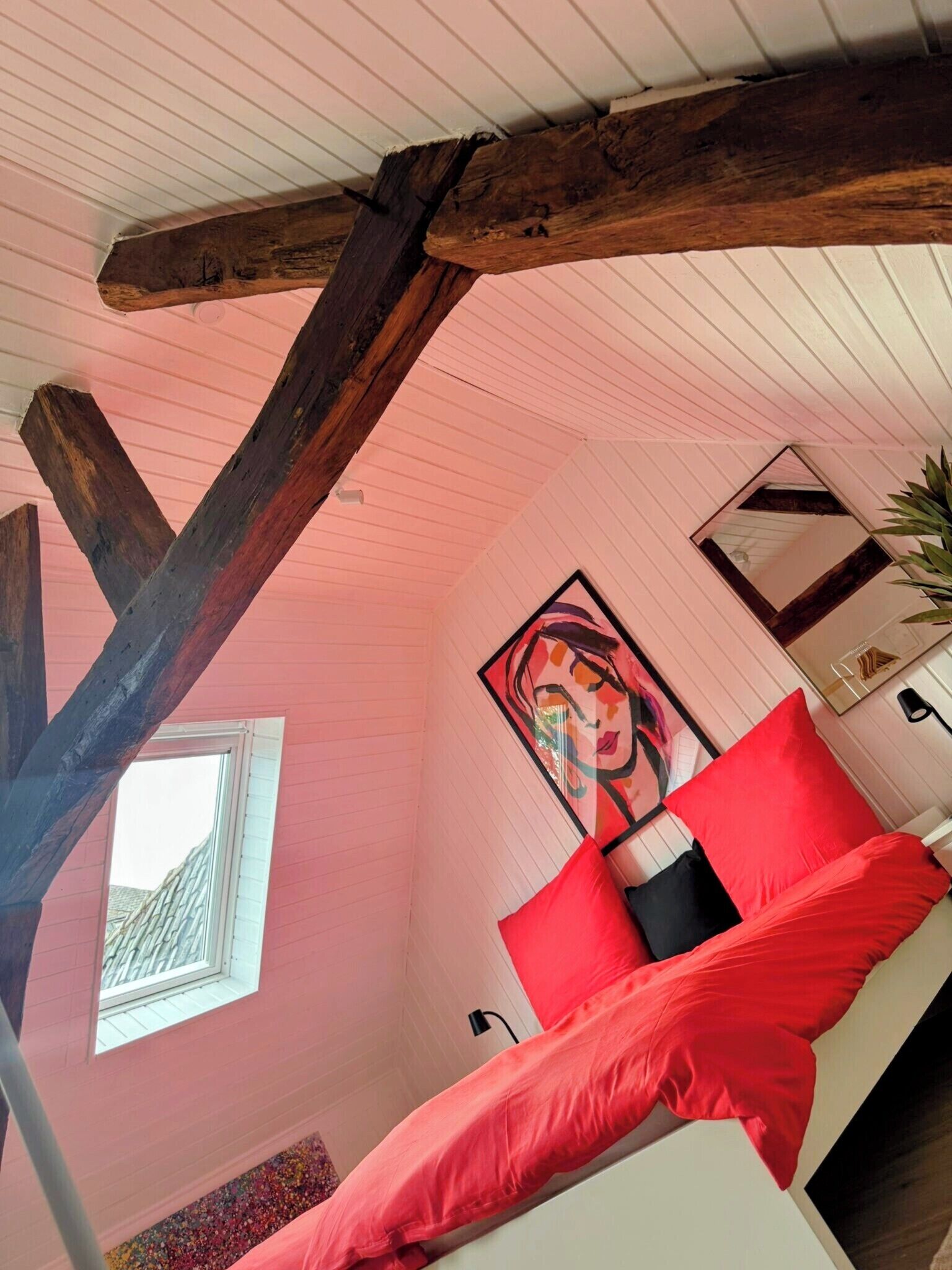 Une pièce avec un plafond en pente, des poutres en bois, un lit avec une literie rouge, une fenêtre et un tableau.
