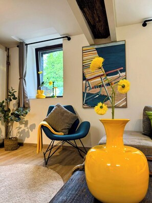 A living room with a large window, a painting, a yellow vase with flowers, a blue chair, and a plant.