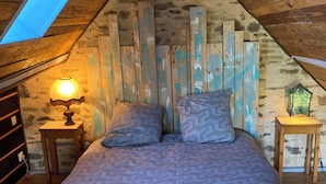 Una camera da letto con soffitto spiovente, pareti in legno, un letto con copriletto decorato, un comodino con una lampada e un piccolo tavolo con una lampada verde.