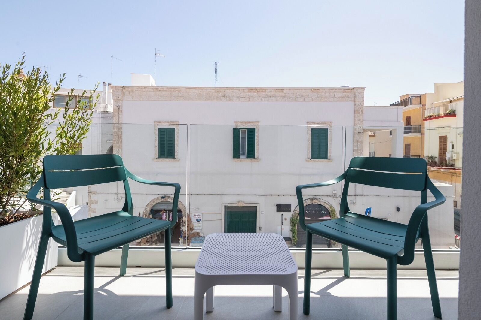 Ein Balkon mit zwei grünen Stühlen und einem kleinen weißen Tisch, Blick auf ein Gebäude mit grünen Fensterläden.