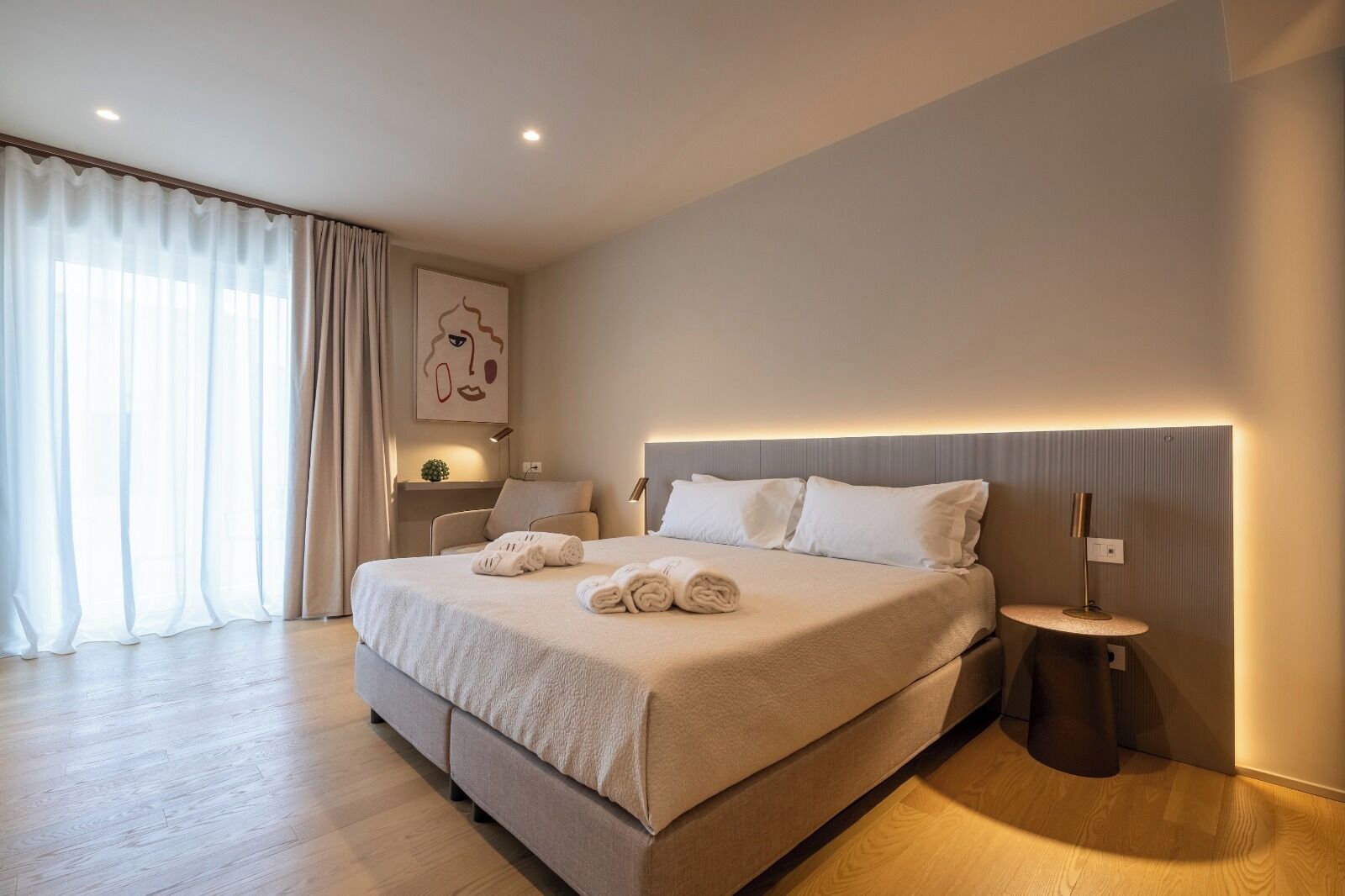 Ein modernes Hotelzimmer mit einem großen Bett, einem Nachttisch, einer Lampe und Blick ins Freie durch transparente Vorhänge.