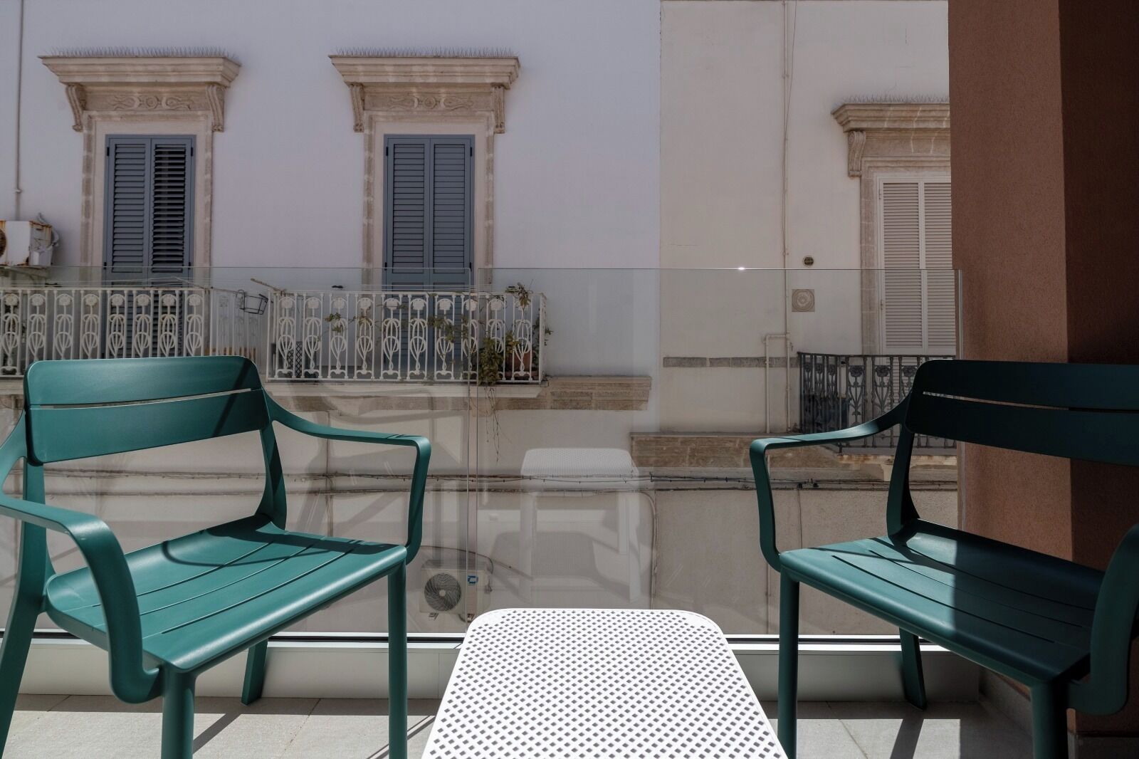 Un balcone con due poltrone verdi e un tavolino, vista su un edificio con finestre e un muro bianco.