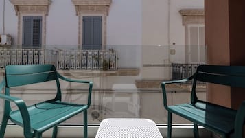 Un balcone con due poltrone verdi e un tavolino, vista su un edificio con finestre e un muro bianco.