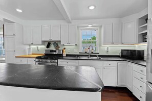 A modern kitchen with white cabinets, a black countertop, stainless steel appliances, and a window providing a view of greenery. - Charming Lakeside Village Farmhouse Retreat (Harrison)