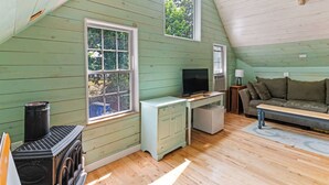 A cozy attic room with a wooden floor, a television, a sofa, and a window overlooking greenery.