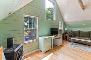 A cozy attic room with a wooden floor, a television, a sofa, and a window overlooking greenery. - Charming Lakeside Village Farmhouse Retreat (Harrison)
