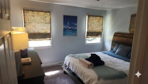 A bedroom with a bed, two windows with curtains, a lamp, and a painting on the wall. - Private 2BR Bungalow • Deck & Grill • Near Newport & RI Coastline  (North Kingstown)