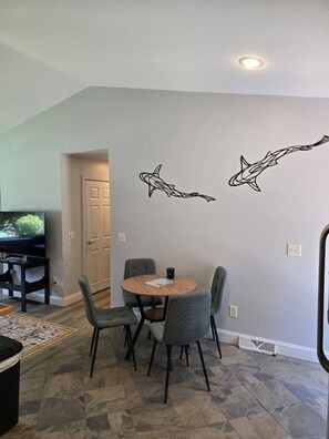 A dining area with a round table and four chairs, a television in the background, and a wall with shark decals. - Private 2BR Bungalow • Deck & Grill • Near Newport & RI Coastline  (North Kingstown)
