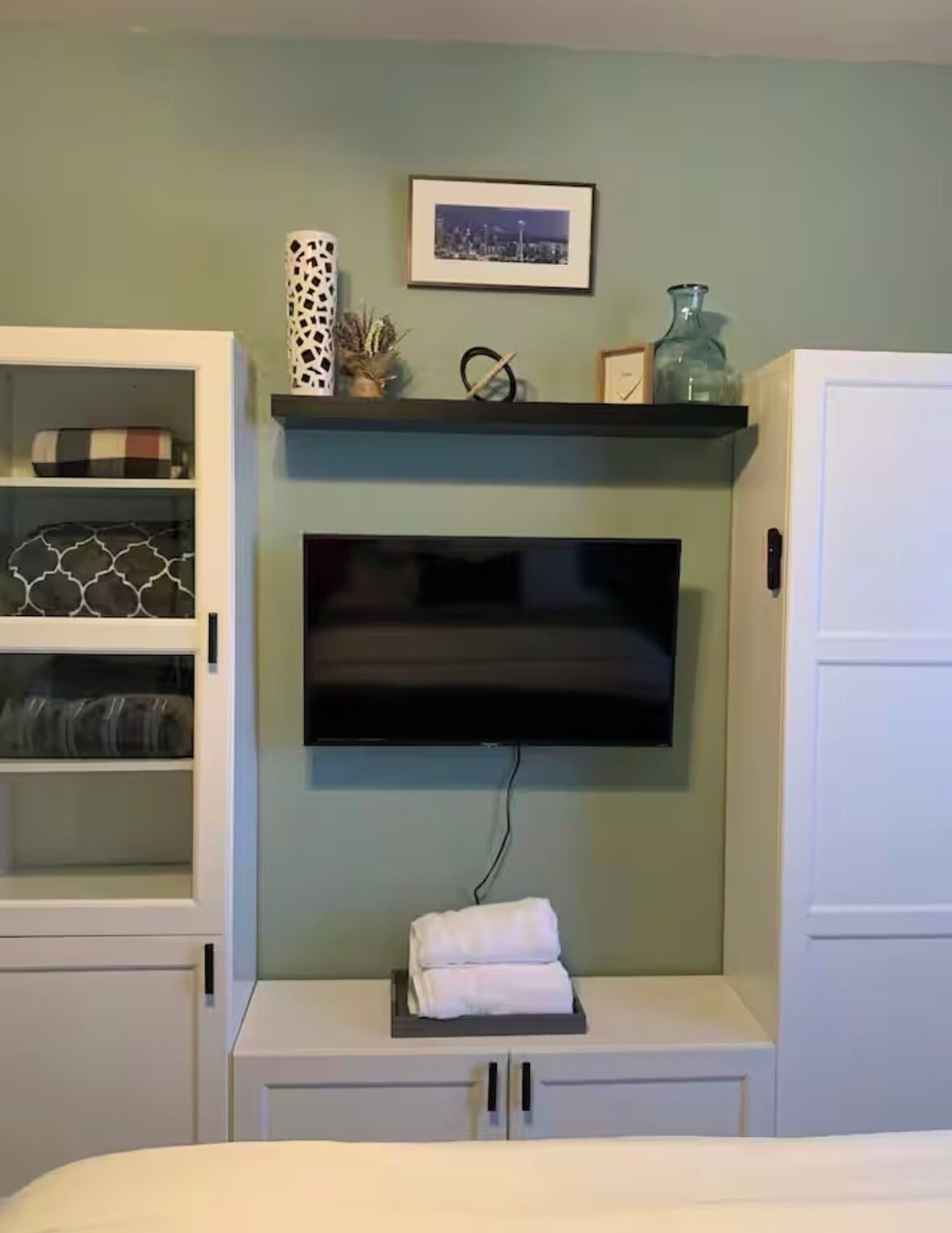 Een kamer met een wandgemonteerde flatscreen televisie, een wit kastje met glazen deuren links, en een witte kast rechts.