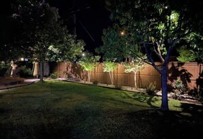 A nighttime view of a well-lit backyard with a wooden fence, trees, and a lawn. - Luxury Pool Oasis  2 primary suites/5 rooms/10 beds. Perfect for multi families (San Antonio)