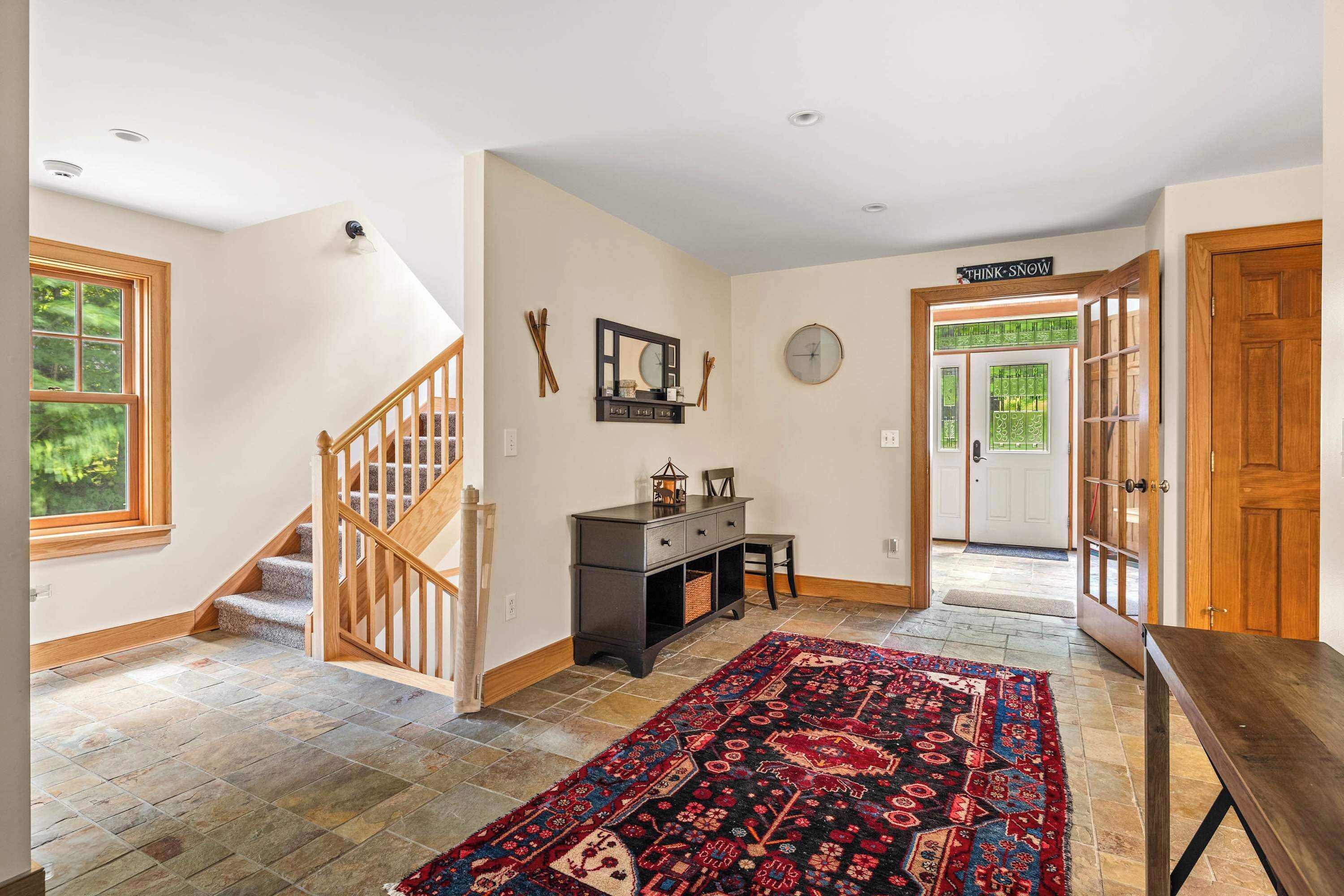A spacious hallway with a wooden staircase, a dark console table, a patterned rug, and a door with a 'Think Snow' sign.