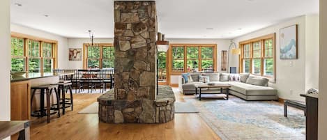 Un salón amplio con chimenea de piedra, suelo de madera, un sofá y ventanas grandes.