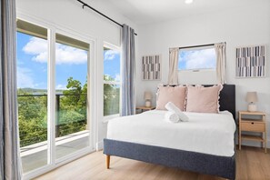A bedroom with a large bed, two bedside tables, a window with curtains, and a view of greenery. - Hilltop Home + Guest Casita w Sweeping Lake Travis Views from Your Rooftop Deck (Austin)