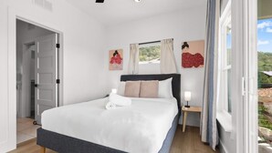 Un dormitorio con cama, mesita de noche con lámpara, ventana con cortinas y una obra de arte en la pared.