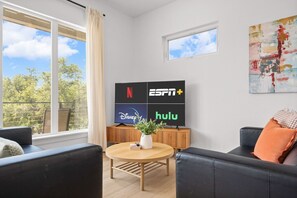A modern living room with a large window, a flat-screen TV displaying streaming services, a round wooden table, and black leather sofas. - Hilltop Home + Guest Casita w Sweeping Lake Travis Views from Your Rooftop Deck (Austin)