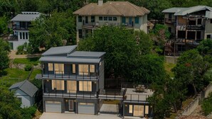 Una casa moderna de dos pisos con garaje, rodeada de exuberante vegetación y otras edificaciones residenciales.