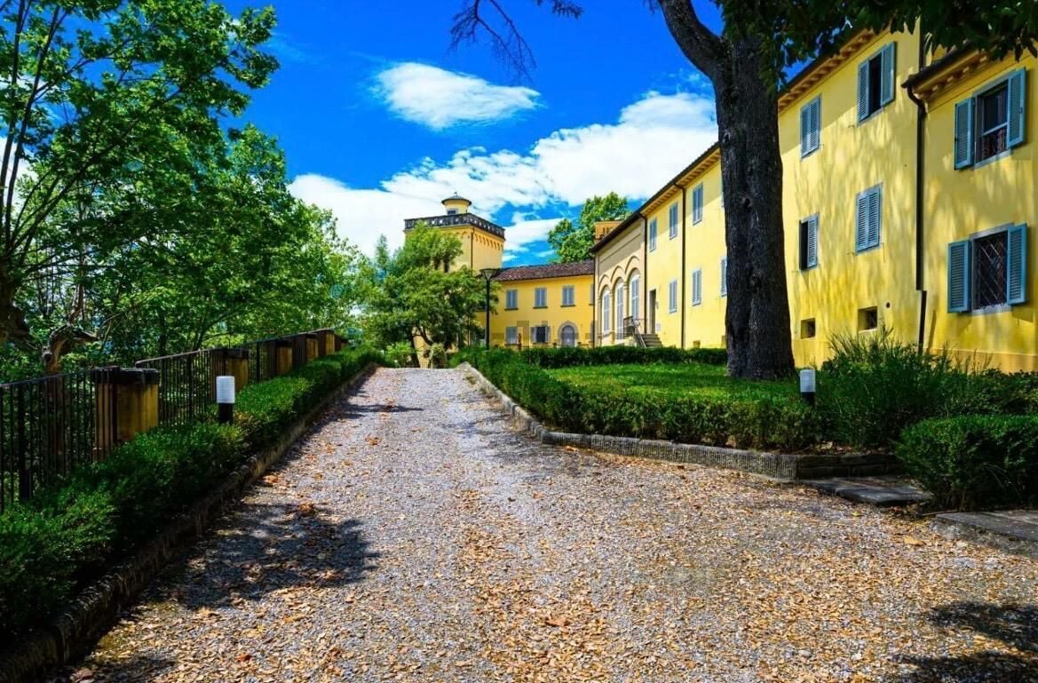 A pathway leading to a yellow building with a tower, surrounded by greenery and a clear sky.