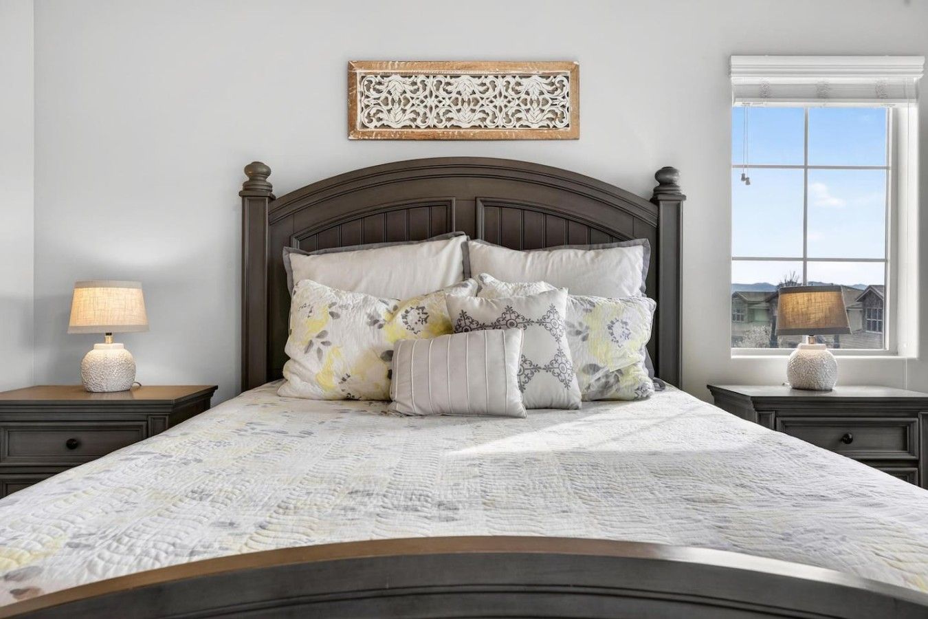 Una camera da letto con una testiera in legno scuro, numerosi cuscini, una lampada da comodino e una finestra vista.