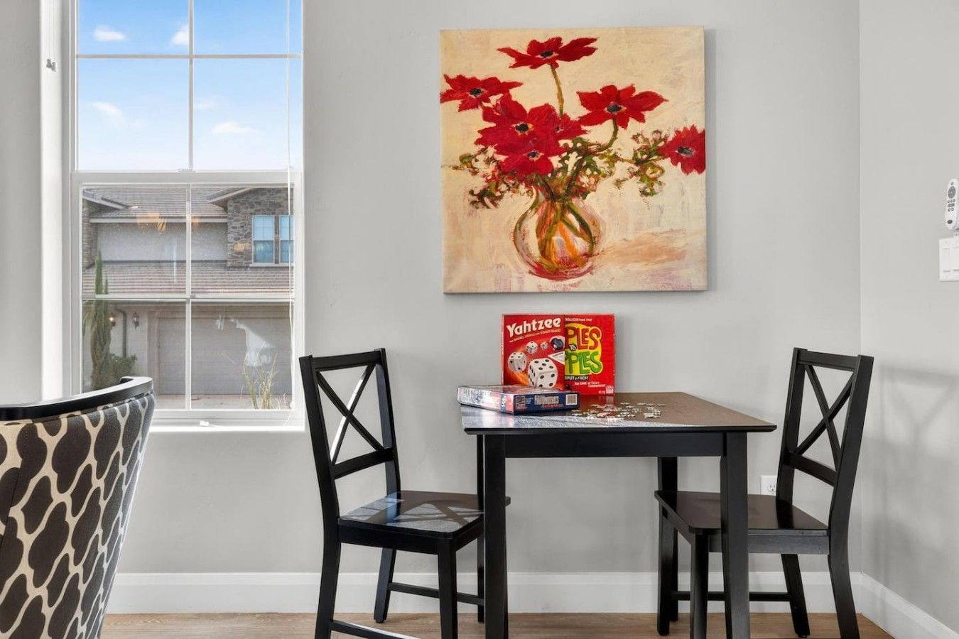 Un coin repas comprenant une table, deux chaises, un tableau représentant des fleurs rouges et un jeu de société.