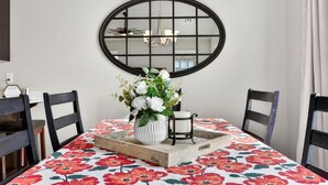 Comedor con una mesa dispuesta para cuatro, mantel con motivos florales, un jarrón blanco con flores y un espejo redondo en la pared.