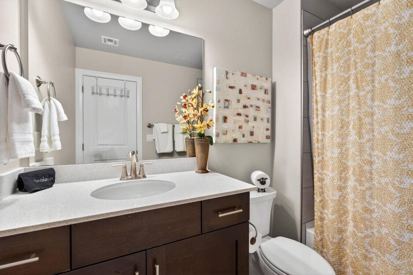 A bathroom with a double sink, a mirror with lights, a toilet, and a shower curtain.