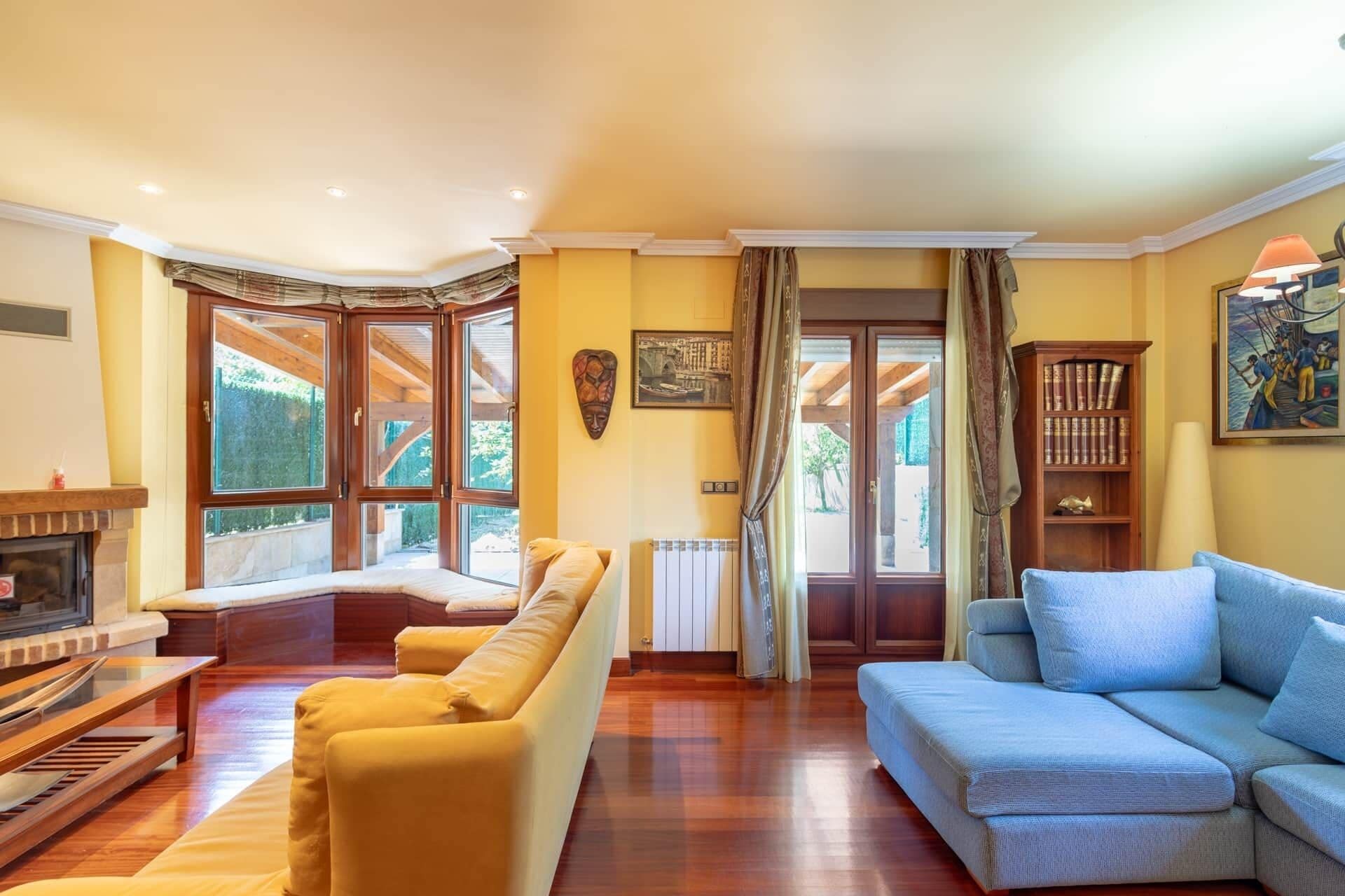 A room with a fireplace, two sofas, a bookshelf, and large windows.