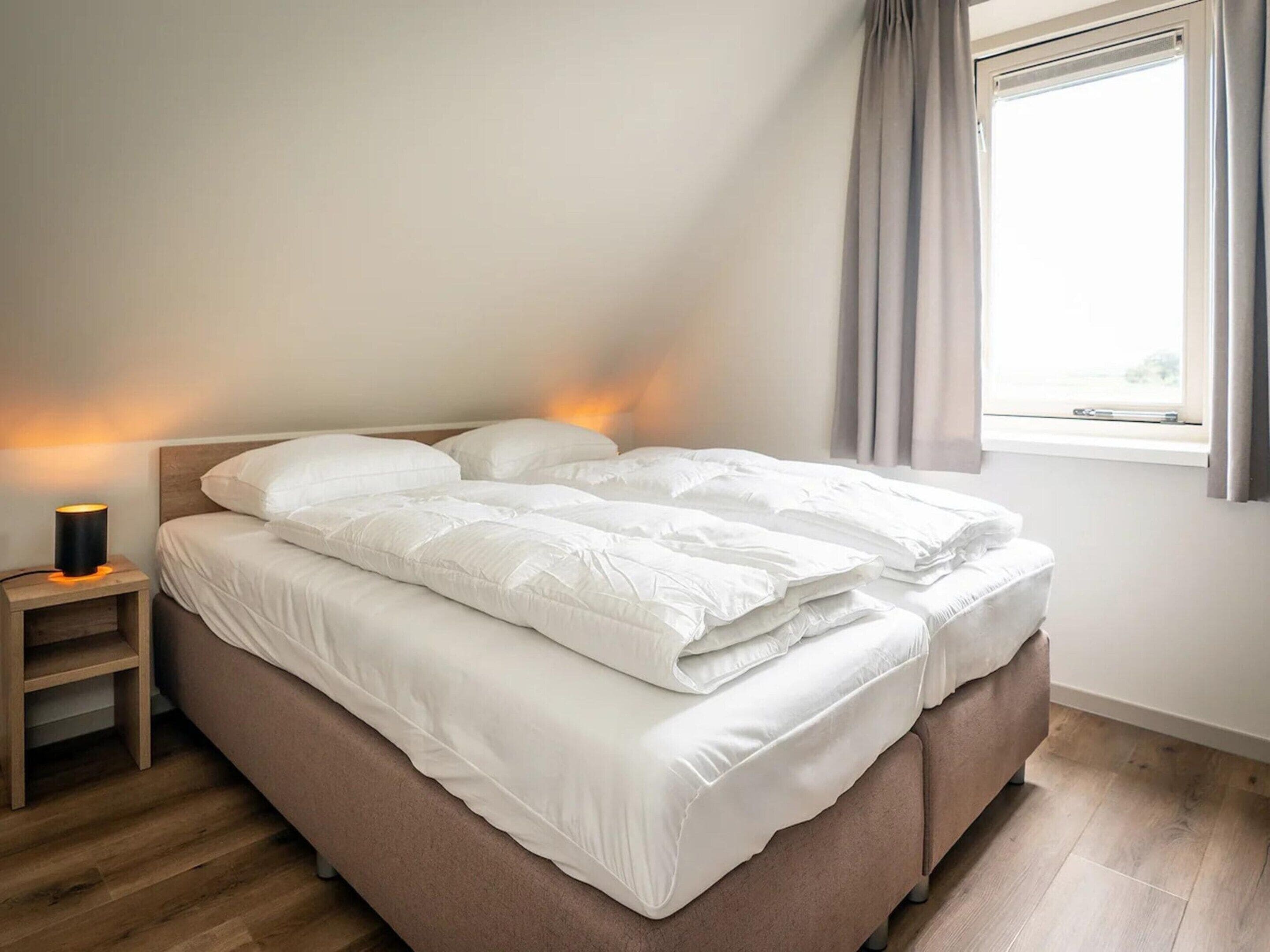 A neatly made bed with white bedding, a bedside lamp, and a window with curtains.