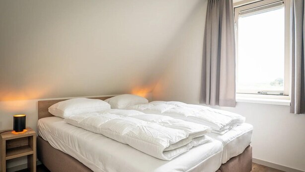 Un letto rifatto con lenzuola bianche, una lampada da comodino e una finestra con tende.