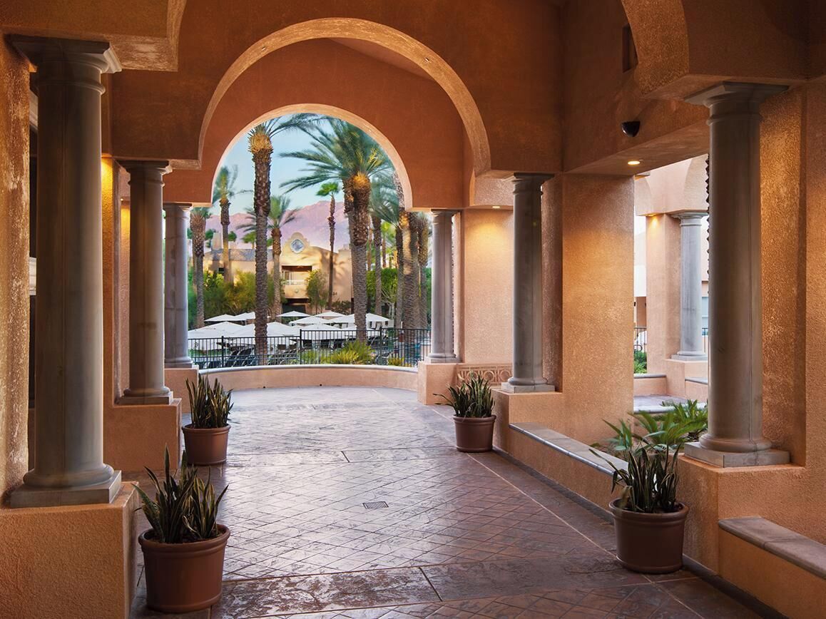 An indoor corridor with columns, potted plants, and a view of outdoor umbrellas.