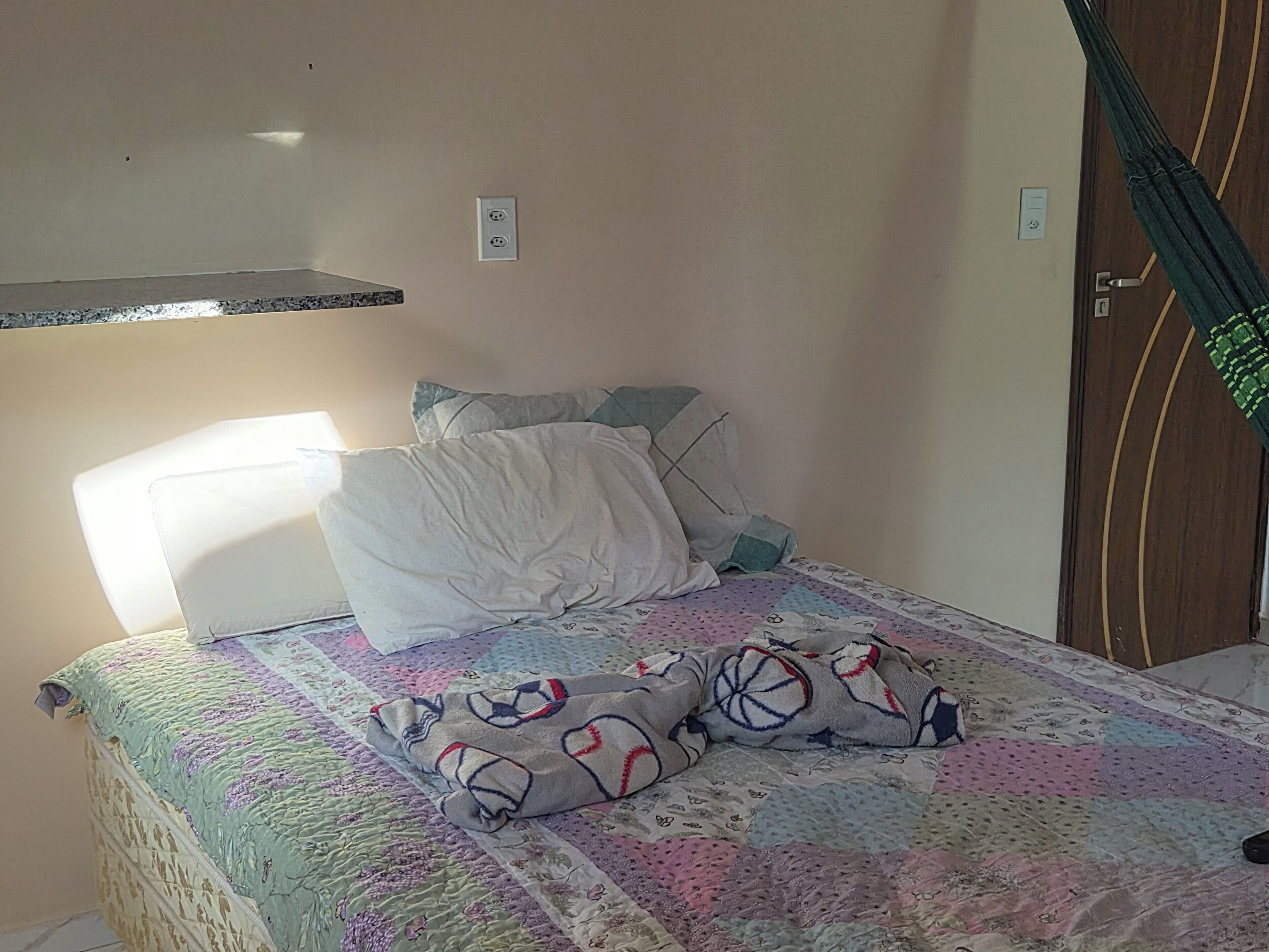 Ein Bett mit einer Decke, Kissen und einem Kopfteil. Eine Steckdose befindet sich an der Wand und eine Tür mit Vorhang ist vorhanden.