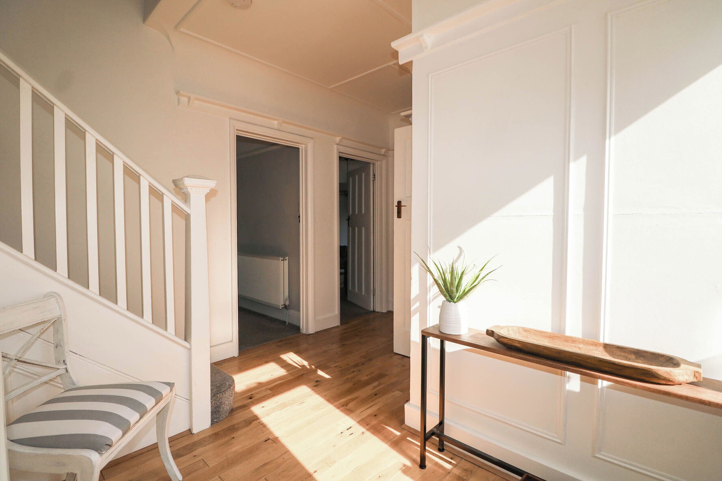 A hallway with a wooden floor, white walls, a white chair, a small table with a plant, and a radiator.