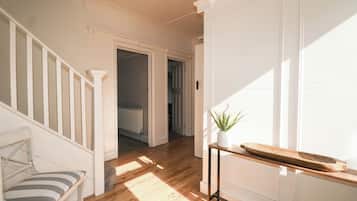 A hallway with a wooden floor, white walls, a white chair, a small table with a plant, and a radiator.