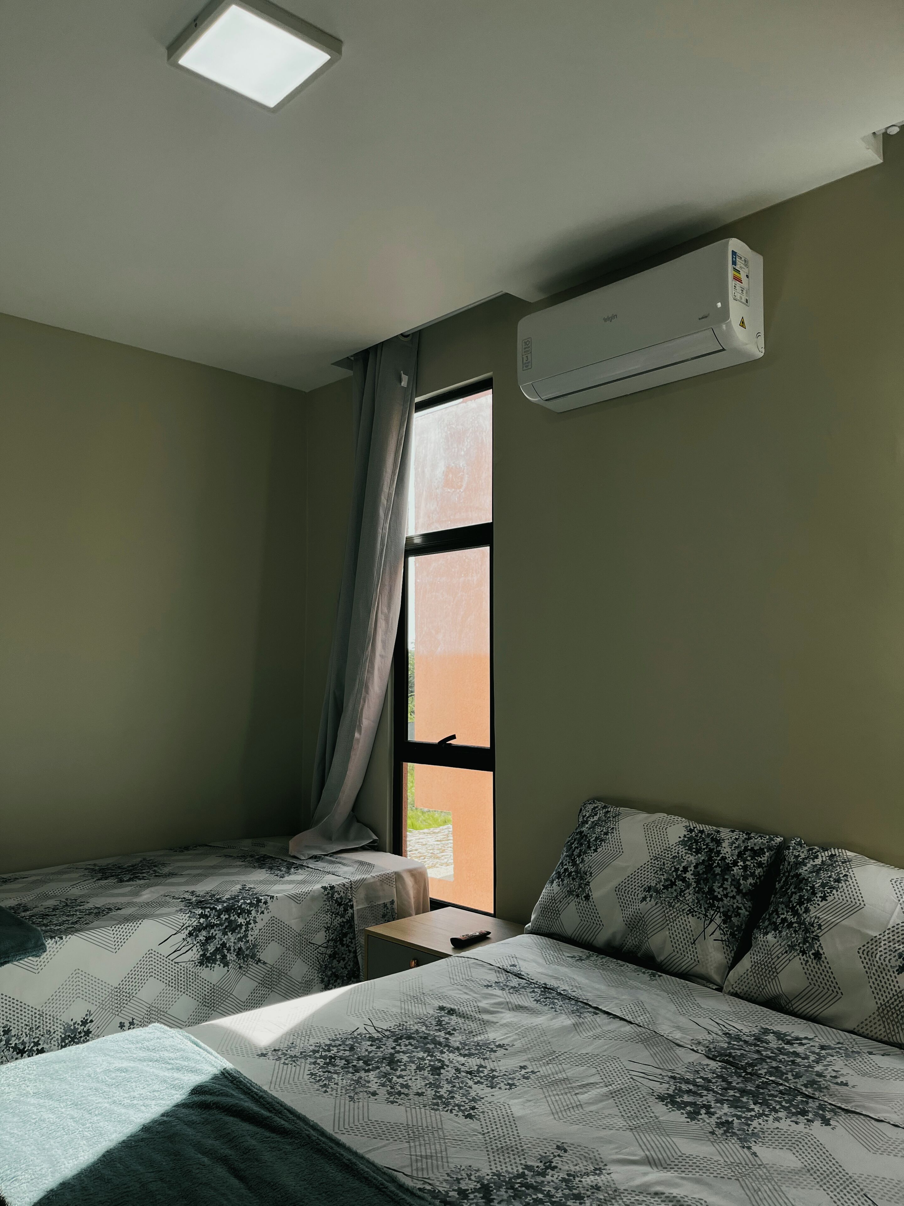 A hotel room with two beds, a window with curtains, an air conditioning unit, and a small table.