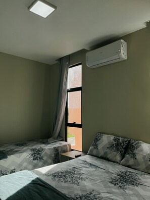 Ett hotellrum med två sängar, ett fönster med gardiner, en luftkonditioneringsanläggning och ett litet bord.