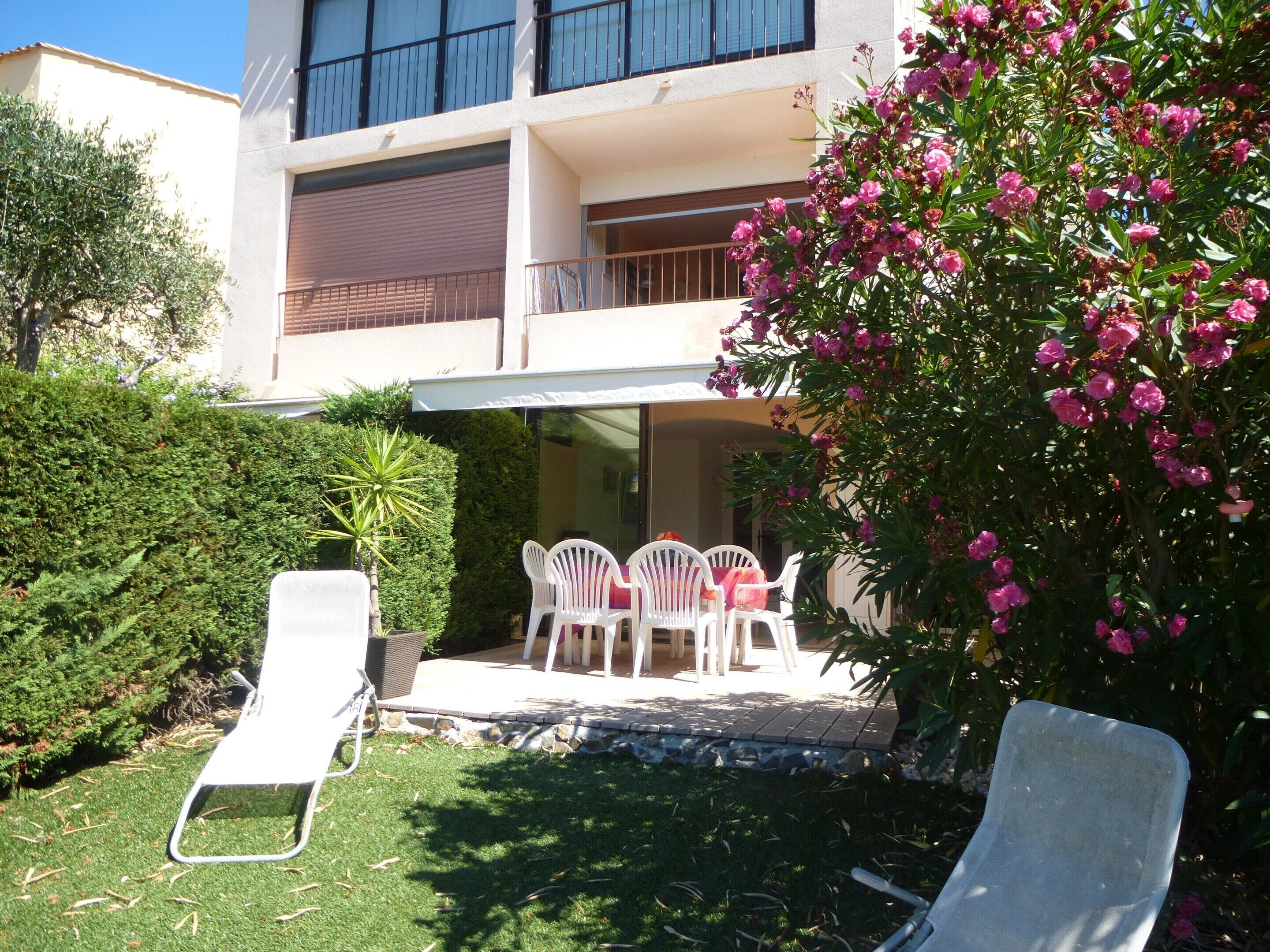 A residential building with a balcony, a garden with chairs, and a flowering bush.