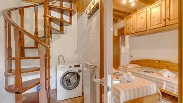 A compact room with a wooden spiral staircase, a washing machine, a bed with a striped bedspread, and wooden cabinets.