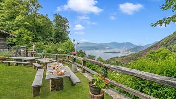 A picnic area with stone tables and benches, surrounded by a wooden fence, overlooking a lake and distant mountains.