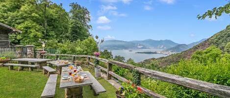 A picnic area with stone tables and benches, surrounded by a wooden fence, overlooking a lake and distant mountains.