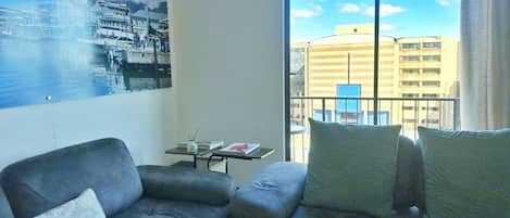 A living room with a sofa, a small table with a book, and a large window with a view of buildings.