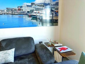Ein Wohnzimmer mit einer Couch, einem kleinen Tisch mit einer Zeitschrift und einem großen Fenster mit Blick auf einen Hafen.