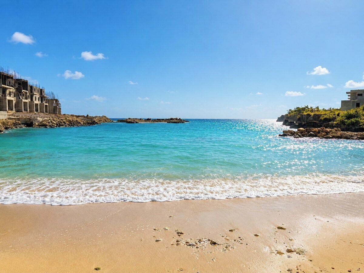 A beach with clear turquoise water, a sandy shore, and buildings on the left.