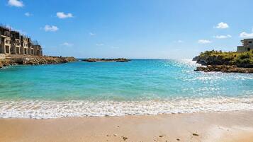A beach with clear turquoise water, a sandy shore, and buildings on the left.