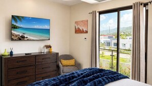 Un dormitorio con cama, cómoda, silla, un cuadro y vistas a edificios a través de una ventana.