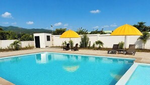A swimming pool with two sun umbrellas, lounge chairs, and a small building in the background. - The Azure Suite Ocho Rios | 2BR (Ocho Rios)