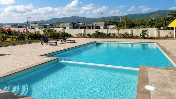 Una piscina rectangular con agua azul cristalina, rodeada por una terraza con baldosas. Hay sillones de descanso y una pequeña mesa en la terraza. Al fondo, se ven edificios y un paisaje montañoso bajo un cielo despejado.