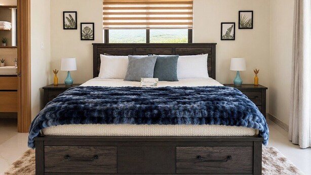 A bedroom with a dark wood bed, white bedding, a fluffy blue blanket, and a window with blinds. - The Azure Suite Ocho Rios | 2BR (Ocho Rios)