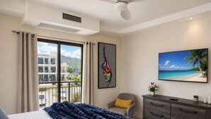 Habitación de hotel con cama, una silla, televisor, cómoda y ventana con vista a un edificio y una playa.