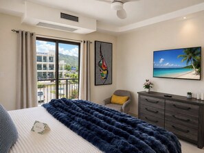 Quarto de hotel com cama, poltrona, televisão, cômoda e janela com vista para um edifício e uma praia.