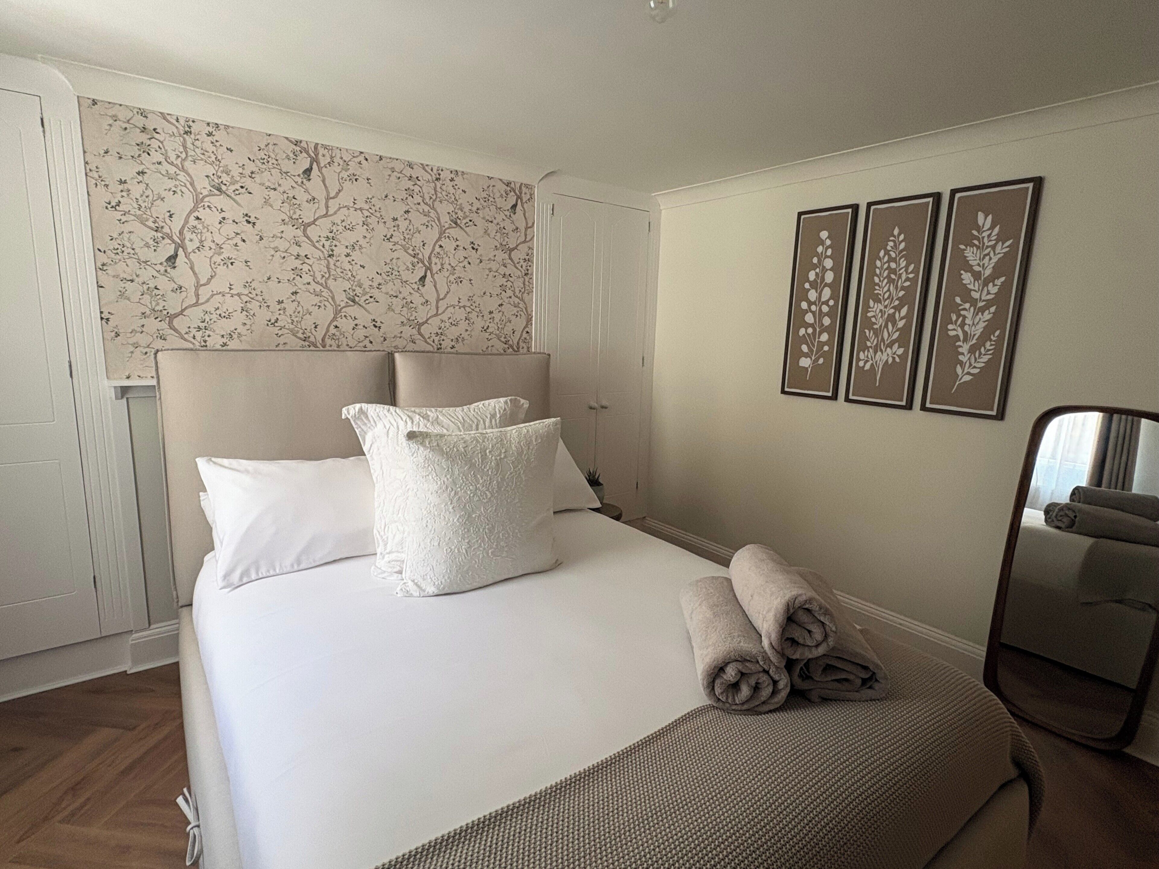 Ein ordentlich bezogenes Bett mit Kissen und aufgerollten Handtüchern, ein Kopfteil und eine Wand mit floralem Tapetenmuster und gerahmten Bildern.